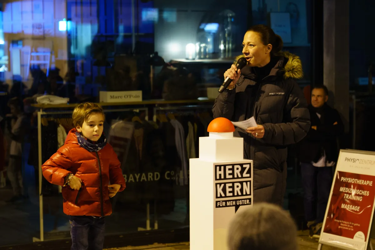 Inszenierte Einschaltung der Weihnachtsbeleuchtung in Uster. Eine Frau spricht in ein Mikrofon, neben ihr steht ein Podest mit einem roten Buzzer, nebendran steht ein Kind.